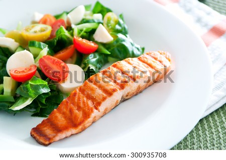 Tasty slice of fried salmon served with mix salad of kale leaves, cherry tomatoes and bocconcini cut in halves, cucumber, served on a white plate. Healthy food. Selective focus, copy space