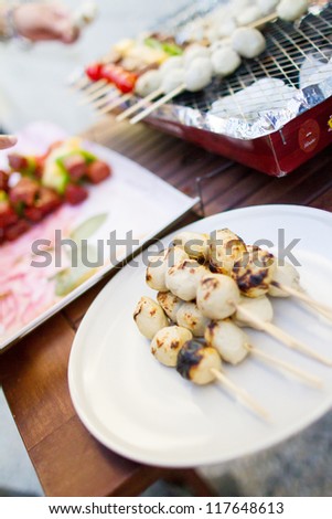 Thai style BBQ chicken balls on skewer grilling on a plate in outdoor cooking area.