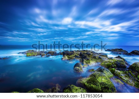Dusk (Sunset) over green moss covered black volcanic rocky outcrop in the middle of the blue ocean with calm sea and sky full of rolling cloud in Port Fairy, Victoria, Australia.