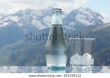 Mineral water bottle and glass with ice cubes, Alps mountains on the background