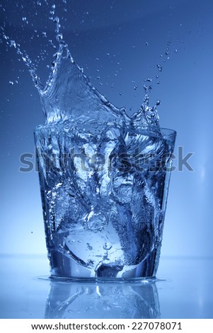 Water glass with a splash of falling ice cube isolated on blue background