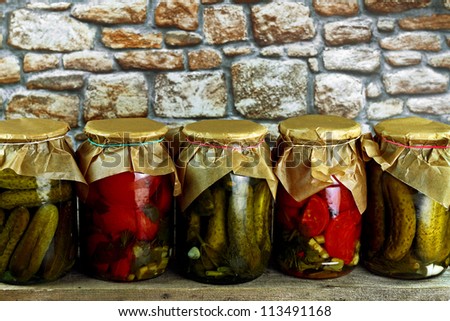 Jars with pickled green cucumbers and red tomatoes on wooden shelf against stone wall
