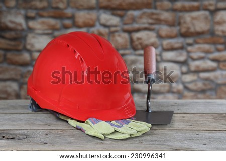 Red protective helmet, gloves and bricklayer trowel lie on rough wooden table on blurred stone wall background. Masonry and building profession concept