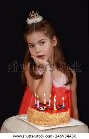 Princess anniversary - cute girl in red dress and crown on head is thoughtful near birthday cake with burning candles - low key portrait on black background