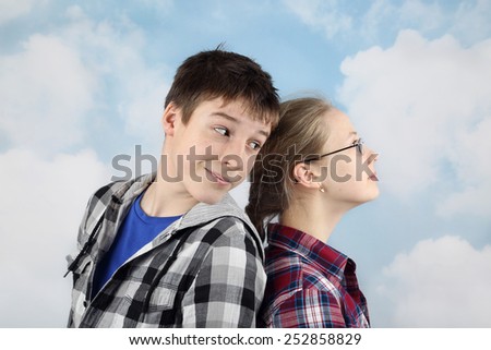 Teenage relationship and friendship - teenagers in plaid shirts stand back to back on cloudy blue sky background. Boy looks at girl with interest
