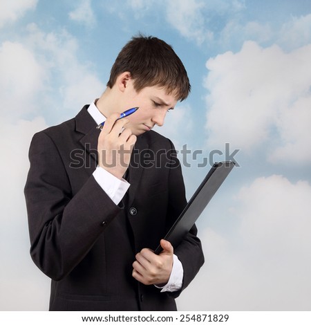 Young manager - Teenage boy in business suit with pen and folder in hands thinks on cloudy blue sky background - side view