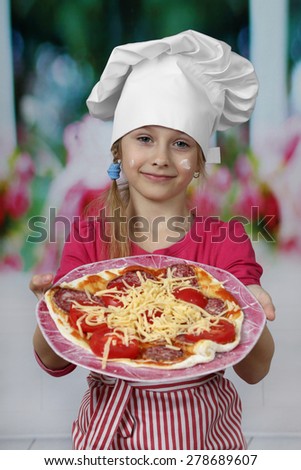 Cute little girl in chief hat shows own hand cooked pizza with sausages and grated cheese - cooking, food and pleasure concept