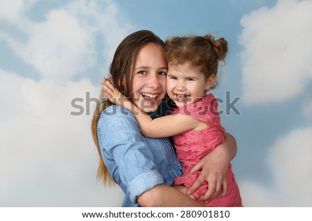 Older girl carries little sister on hands on cloudy blue sky background - Family, childcare and love concept