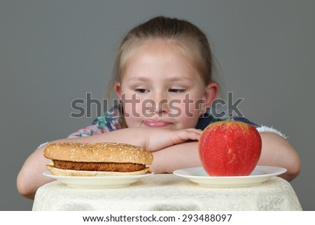 Cute little girl makes choice between hamburger and apple on gray background - Fast food, diet and healthy eating concept