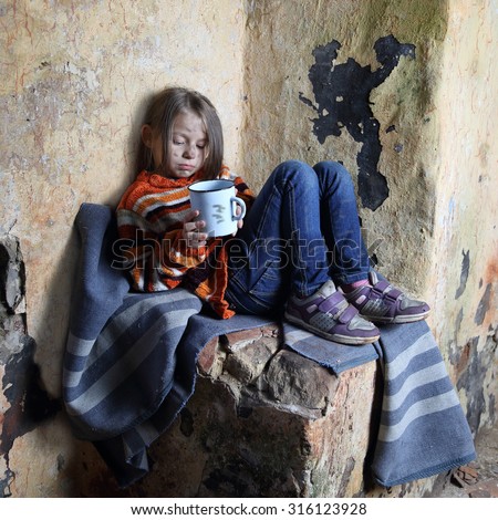 Sad little girl with dirty face sits on old blanket in basement with iron mug in hands - refugee orphan, poverty concept