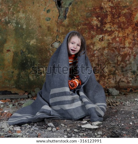 Little girl sits in basement wrapped in blanket and crying with tears on face - orphan, poverty, despair concept