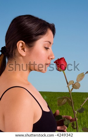 A beautiful woman smelling a red rose on a green grassy field with a blue sky background