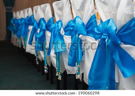 Ranged in a line wedding chairs with white covers and blue marine bow and small shells on the end of a bow