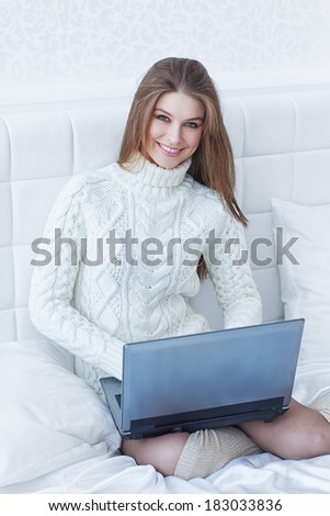 beautiful smiling young woman in white sweater sitting in the bed using laptop
