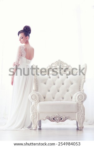 Full-length portrait of young gorgeous bride standing near antique armchair