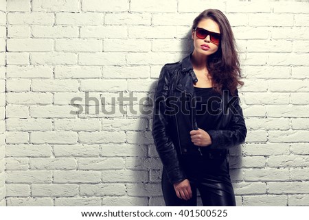 fashion model in sunglasses, black leather jacket, leather pants. Posing near white brick wall.
