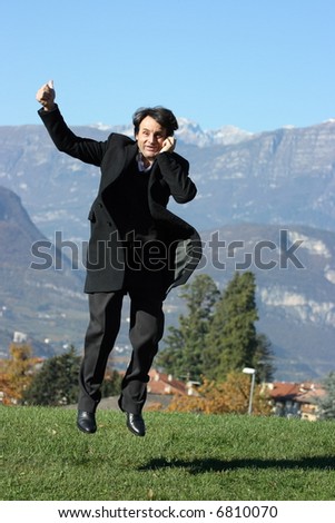 A businessman has just received good news by phone and he is jumping happily because of this. There are Alps on the background, rural landscape.