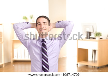 An attractive young executive relaxing at work in an office leaning back with his hands holding his head smiling at camera.