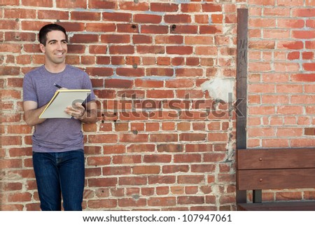 Good looking young adult man writing on a yellow pad of paper by a brick wall outdoors wearing a purple shirt.