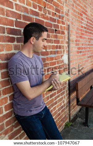 Good looking young adult man writing on a yellow pad of paper by a brick wall outdoors wearing a purple shirt.