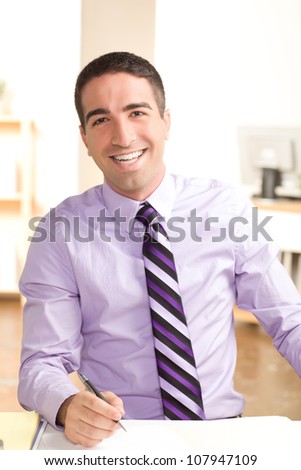 A handsome young man at work with a big smile looking at camera with a pen wearing purple shirt and tie