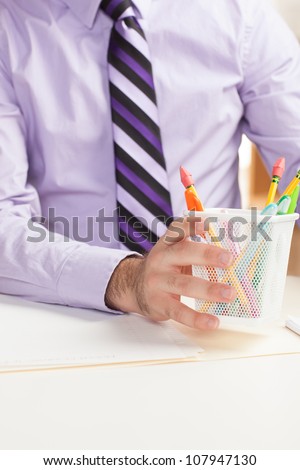 A detail shot of a man at work holding a white pencil cup filled with pens and pencils includes partial torso and purple shirt with tie