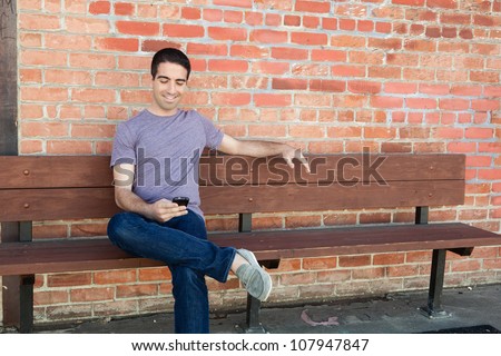 An attractive young man sitting on a bench outside using his cell phone wearing a purple shirt and jeans.
