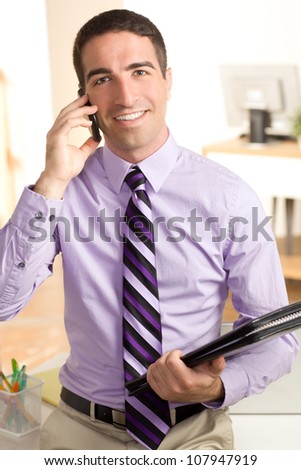 A handsome business man holding a leather folio, talking on cell phone, looking at camera with smile.