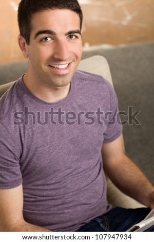 Cute young man sitting in a beige chair reading a newspaper smiling and looking at camera.