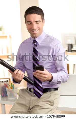 A cute man looking at a smart phone holding a leather pad in an office setting with a big smile on his face