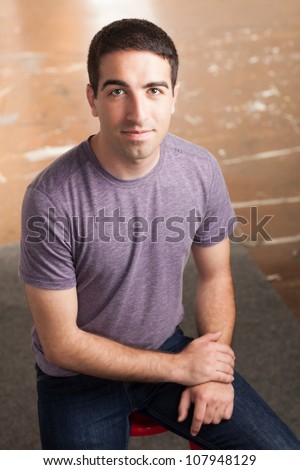 Good looking gentleman sitting on stool wearing purple shirt and jeans looking at camera.