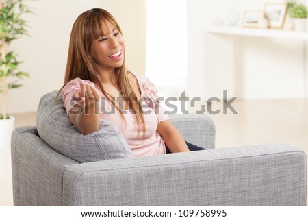 Attractive and vibrant Filipino female relaxing in living room in a home wearing a pink shirt and blue jeans on a grey chair.