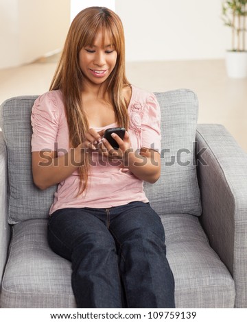 Attractive and vibrant Filipino female relaxing in living room in a home wearing a pink shirt and blue jeans on a grey chair.