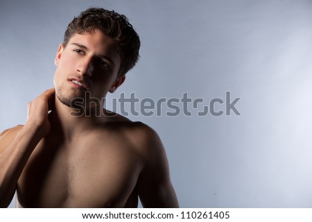 Sexy young guy with shirt off showing abdominal and arm muscles in studio against a grey background.