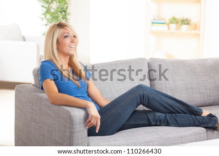 Beautiful Caucasian woman with blond hair relaxing on couch in living room wearing a blue shirt and jeans.