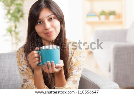 Attractive young ethnic woman in living room holding blue coffee mug wearing festive yellow shirt with flowers.