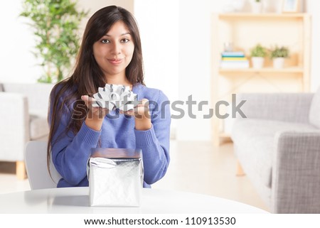 Adorable Hispanic woman wrapping gift with silver wrapping paper and silver bow in living room at table wearing blue shirt.
