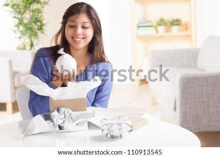 Adorable Hispanic woman opening gift with silver wrapping paper and silver bow in living room at table wearing blue shirt.