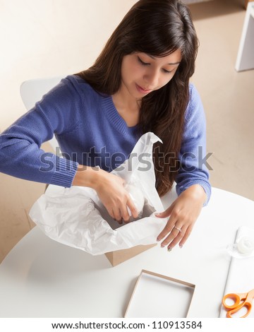 Adorable Hispanic woman wrapping gift with silver wrapping paper and brown box  in living room at table wearing blue shirt.
