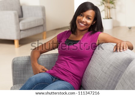 Beautiful youthful African American woman relaxing at home on grey couch wearing pink shirt.