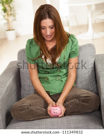 Attracitve young female sitting on chair at home with cute pink piggy bank.