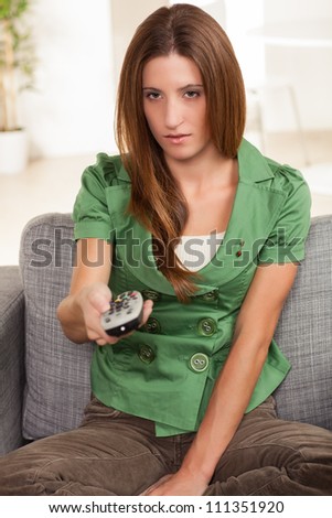 Beautiful female sitting on chair watching television holding a remote control wearing a green shirt and brown pants.