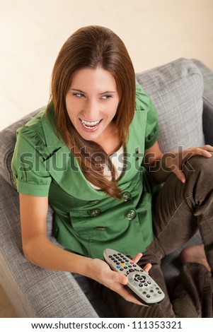 Beautiful female sitting on chair watching television holding a remote control wearing a green shirt and brown pants.