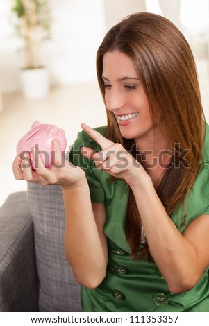 Attractive young female sitting on chair at home with cute pink piggy bank.