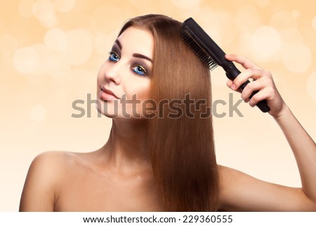 Attractive adult Caucasian girl with blue eyes combing her perfect brown healthy straight hair with comb in studio on abstract yellow background
