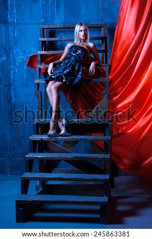 great girl with white hair sitting on stairs and looking to the side . Photo in cold colors with red cloth on background. Studio shot