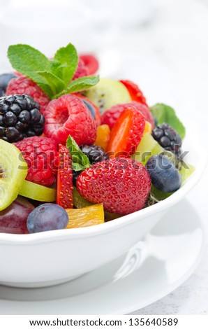 salad of fresh fruit and berries in a white bowl, berries and a cup of tea in the background, closeup vertical