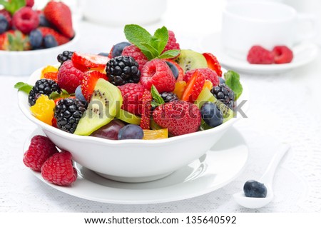 salad of fresh fruit and berries in a white bowl, berries and a cup of tea in the background, horizontal closeup