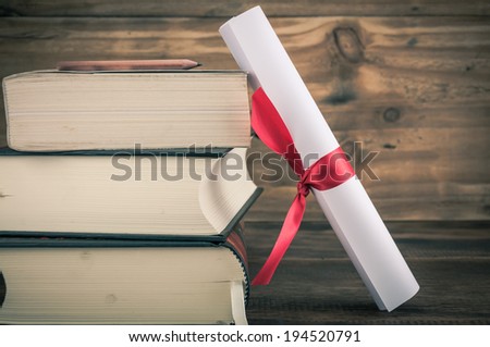 A parchment diploma scroll, rolled up with red ribbon beside a stack of books on wood background with vintage filter