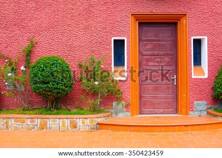 Red door with pink wall and small Bush, with two small windows beside the door and orange floor.